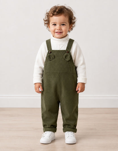 A toddler standing indoors wearing olive green corduroy overall-style pants over a white ribbed turtleneck sweater, with white sneakers, against a plain light wall and wood floor.
