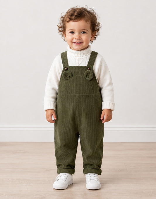 A toddler standing indoors wearing olive green corduroy overall-style pants over a white ribbed turtleneck sweater, with white sneakers, against a plain light wall and wood floor.