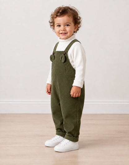 Side view of a toddler standing indoors wearing olive green corduroy overall-style pants over a white ribbed turtleneck sweater, with white sneakers, against a plain light wall and wood floor.