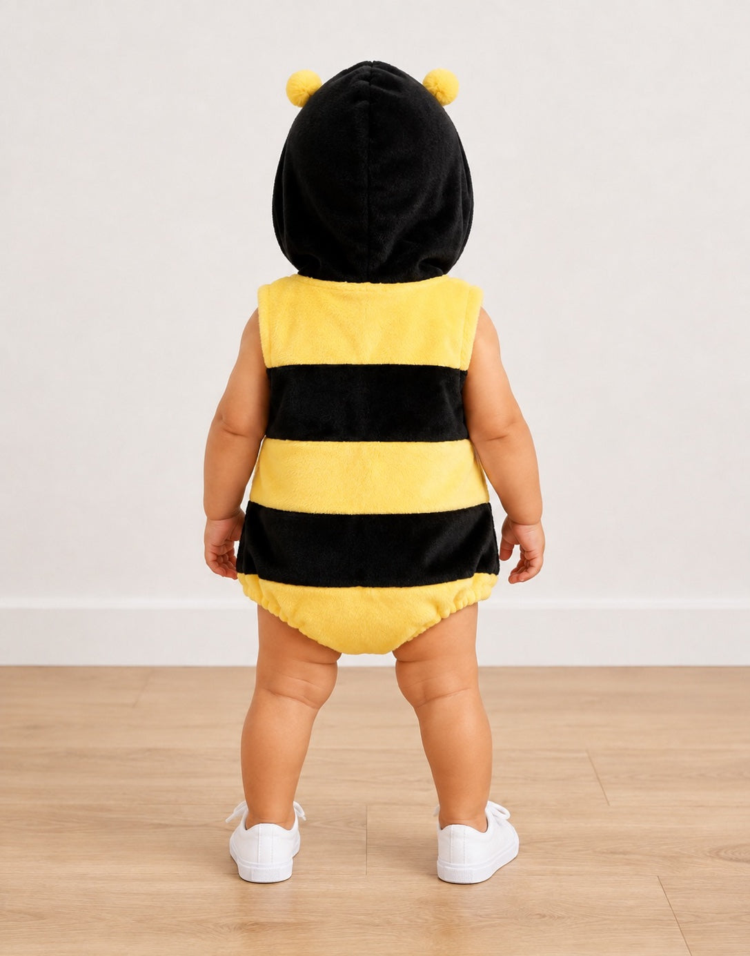 A baby wearing a plush bumblebee costume, viewed from behind. The costume features a black hood with two yellow pom-poms, yellow and black striped body panels, and yellow diaper cover, paired with white shoes, set against a neutral indoor background.