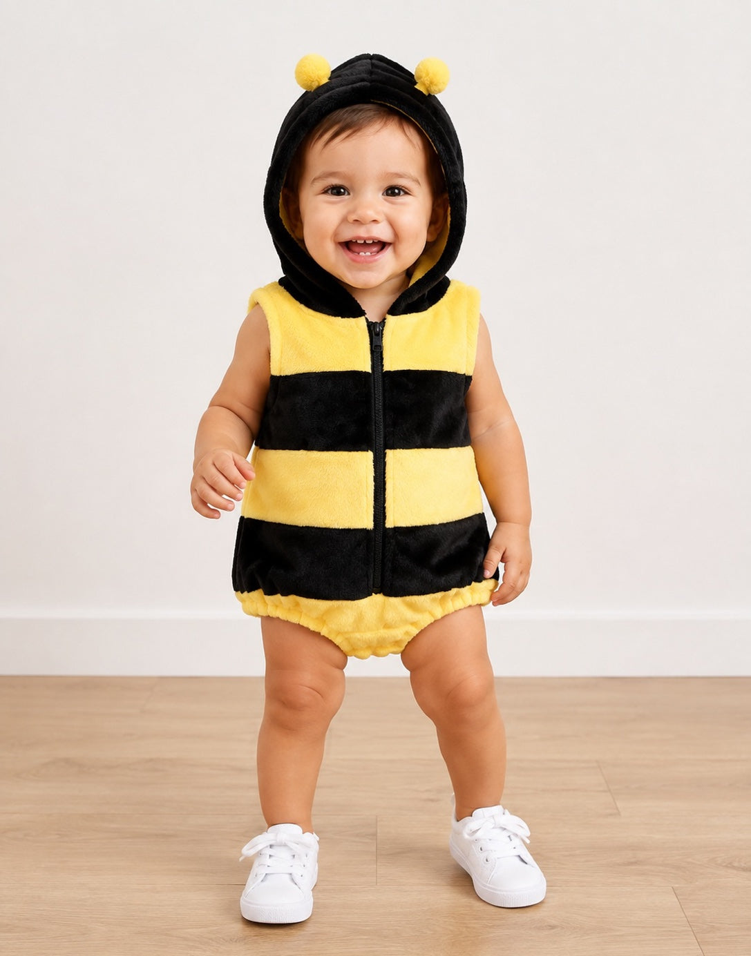 A toddler wearing a yellow and black striped bumblebee costume with a hood featuring two yellow pom-poms, standing on a light wood floor against a white wall.