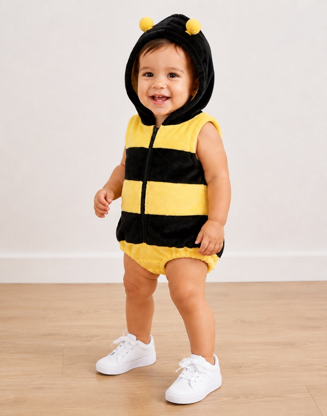 Side view of a baby wearing a plush yellow and black bumble bee costume with a hood featuring yellow pom-poms, standing on a light wood floor against a white wall.