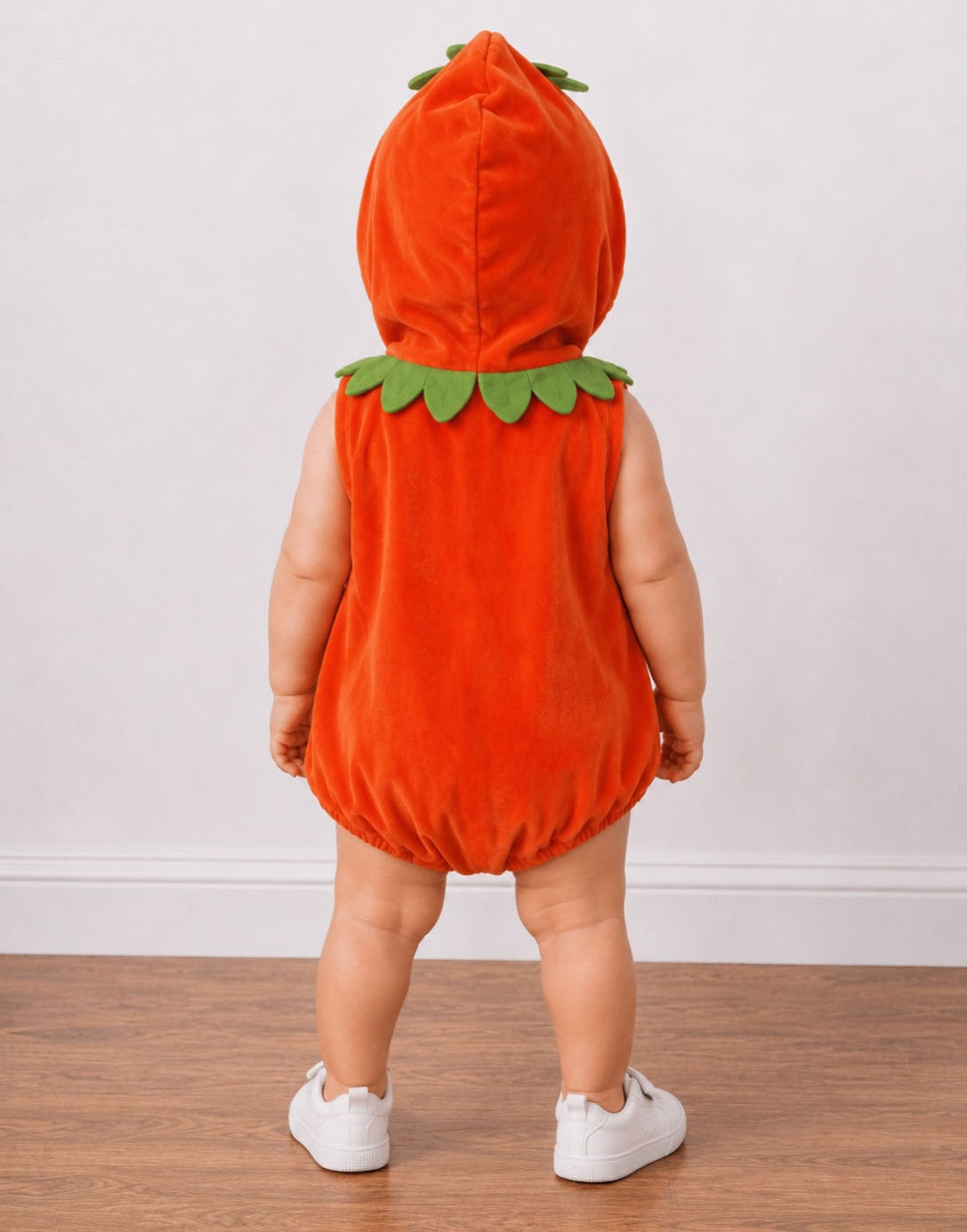 A toddler viewed from behind wearing a bright orange pumpkin costume with green felt leaves around the neck and hood, standing on a wood floor against a plain light wall.