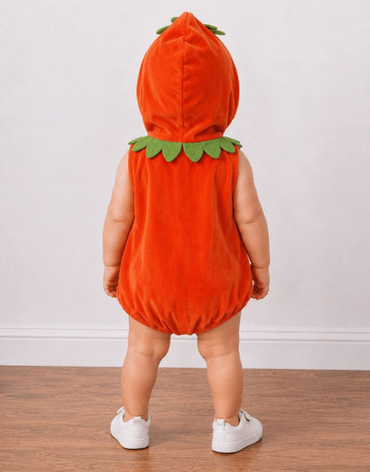 A toddler viewed from behind wearing a bright orange pumpkin costume with green felt leaves around the neck and hood, standing on a wood floor against a plain light wall.