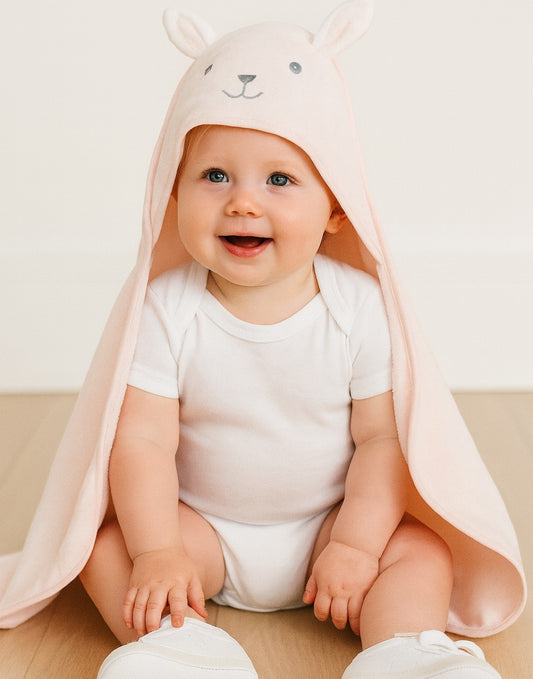 Baby wearing a pink bear hooded towel on a light wooden floor.