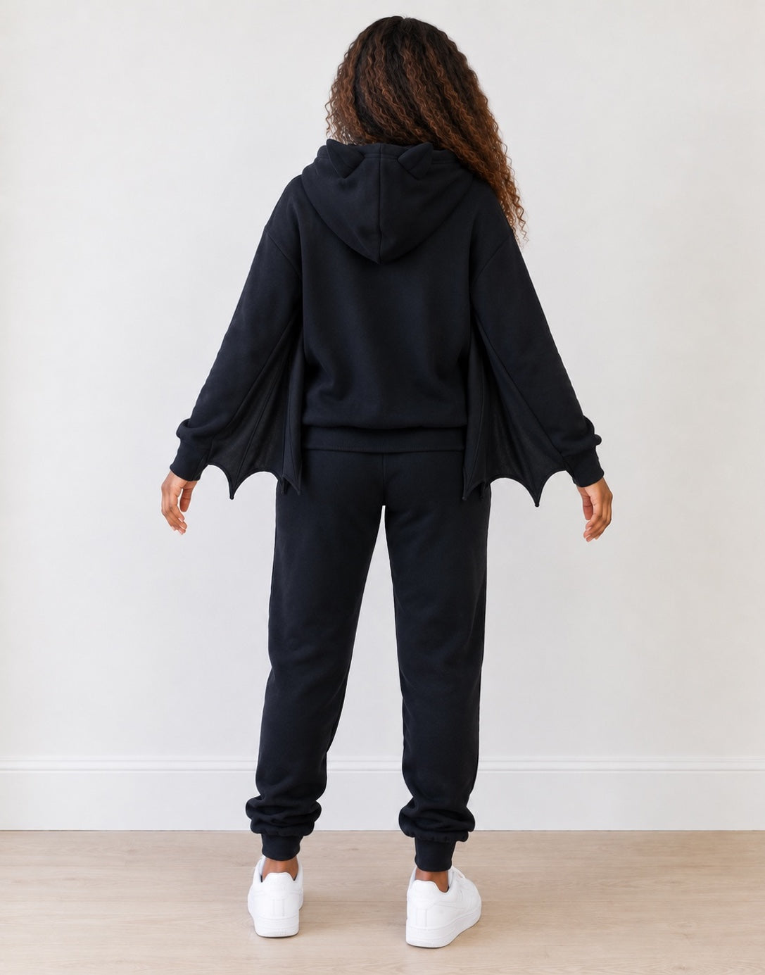 A behind view of a woman wearing a black hoodie with batwings attached to the sleeves, and black joggers with white tennis shoes.