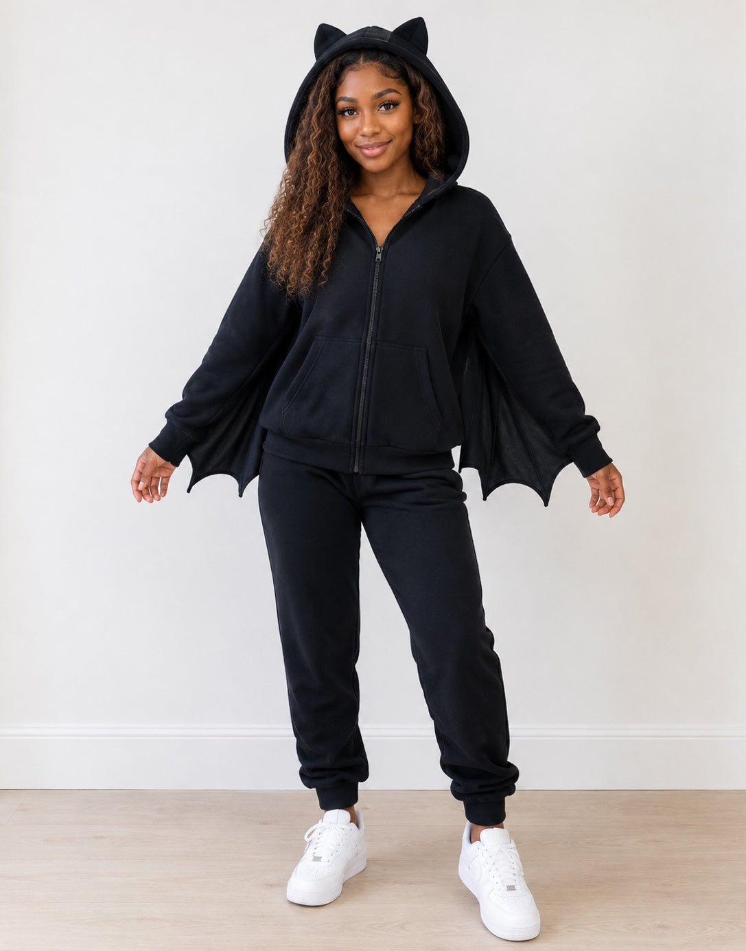 Woman wearing a black hoodie with ears and bat wings attached to the sleeves and black sweatpants with white shoes on a plain background.