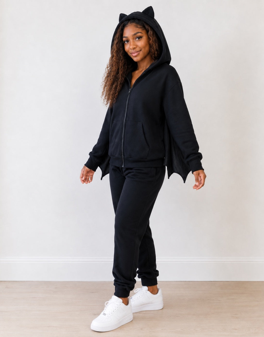 Side view of woman wearing a hooded bat wings costume with black sweatpants and white sneakers on a plain background.