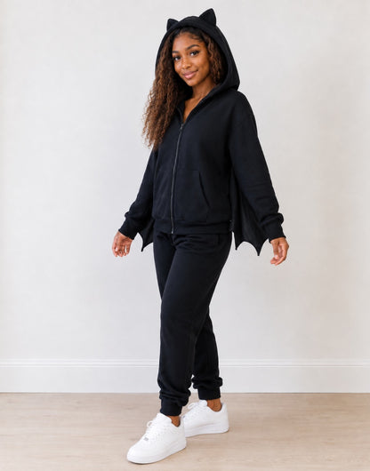 Side view of woman wearing a hooded bat wings costume with black sweatpants and white sneakers on a plain background.