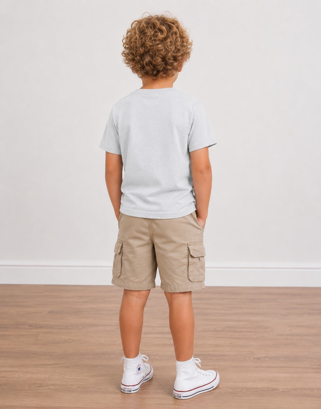 A boy model shown from the back wearing a light gray Crewneck T-Shirt, beige cargo shorts, white socks, and white high-top sneakers, standing on a light wood floor against a white wall.