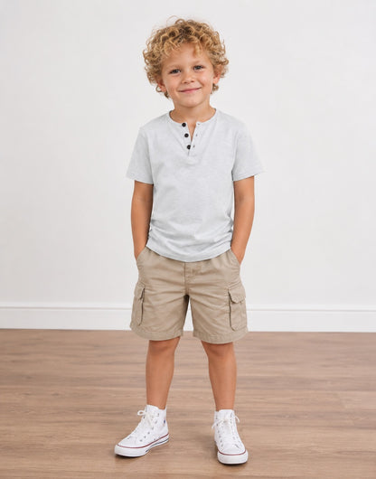 A young boy with curly blonde hair stands in a studio wearing a light gray Button Short Sleeve Front Henley T-Shirt, khaki cargo shorts, and white sneakers against a white wall and wood floor.