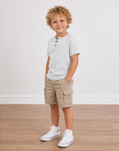 A young boy with curly blonde hair stands in a studio wearing a light gray Short-Sleeve Henley Tee Shirt, khaki cargo shorts, and white sneakers against a white wall and wood floor.