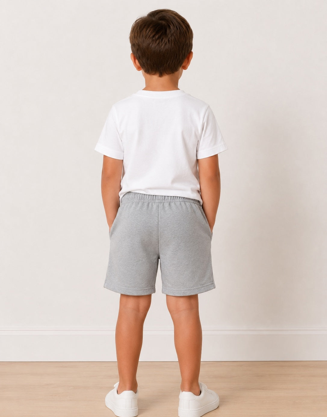 Back view of a young boy stands in a studio wearing a white crewneck t-shirt, light gray heathered drawstring shorts, and white sneakers against a light beige wall and wood floor.