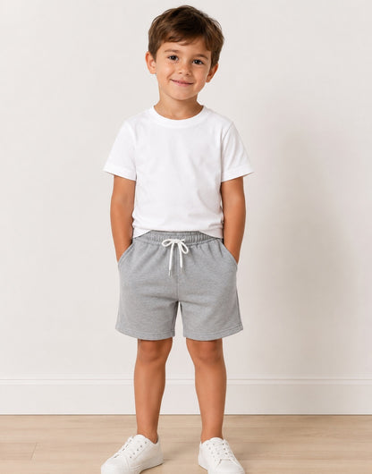 A young boy stands in a studio wearing a white crewneck t-shirt, light gray heathered drawstring shorts, and white sneakers against a light beige wall and wood floor.