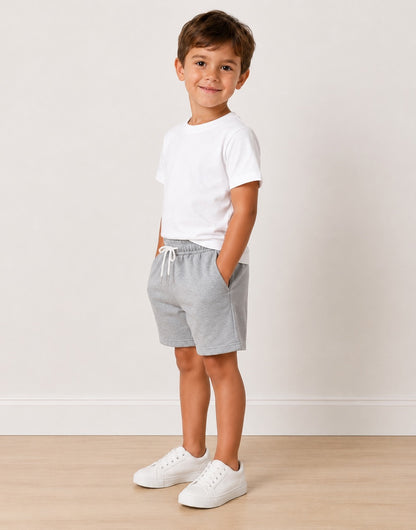 Side view of a young boy stands in a studio wearing a white crewneck t-shirt, light gray heathered drawstring shorts, and white sneakers against a light beige wall and wood floor.