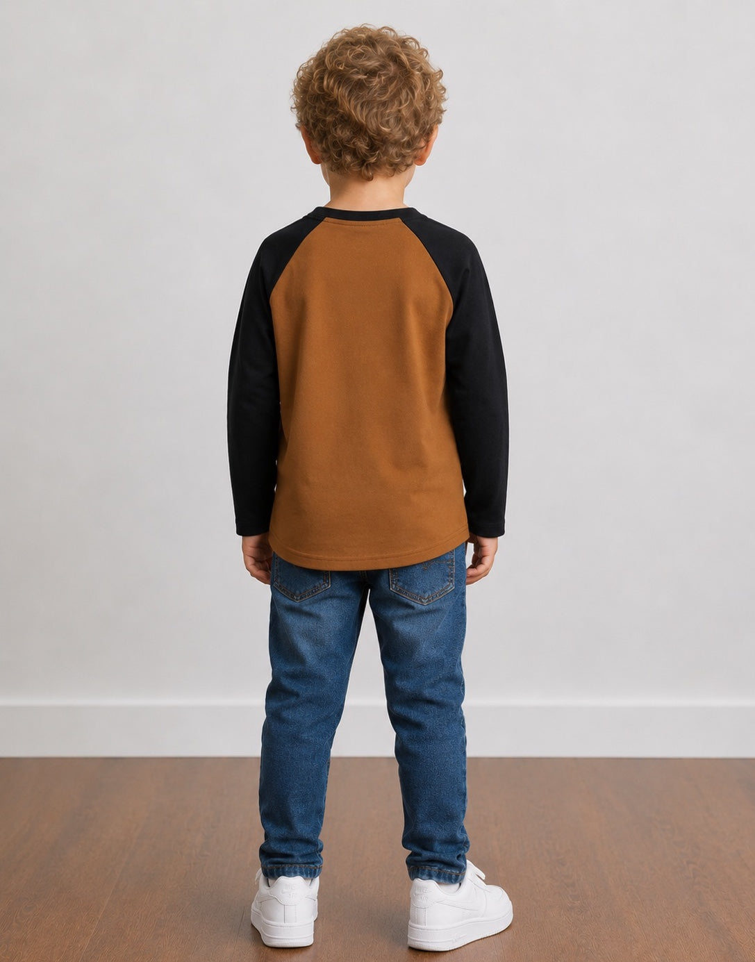 A young boy standing with their back to the camera, wearing a two-tone Raglan Henley Long-Sleeve Shirt with a brown body and black sleeves, blue jeans, and white sneakers, set against a plain light gray wall and wooden floor.