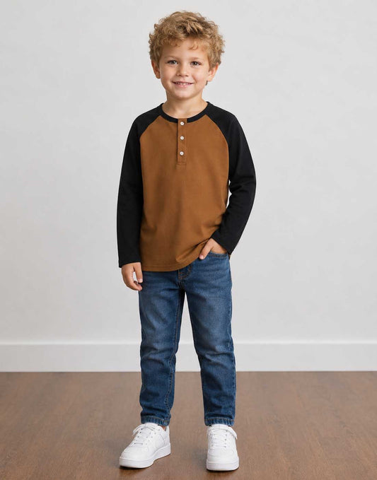 A young boy with curly blond hair stands wearing a brown and black Raglan Henley Long Sleeve Shirt, blue jeans, and white sneakers against a plain light gray wall and wood floor.