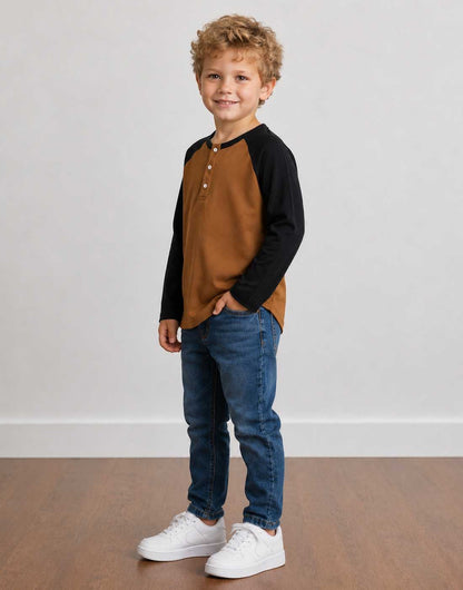 Side view of a young boy with curly blond hair stands in a studio wearing a brown and black Raglan Long Sleeve Henley Top, blue jeans, and white sneakers against a light gray wall and wood floor.