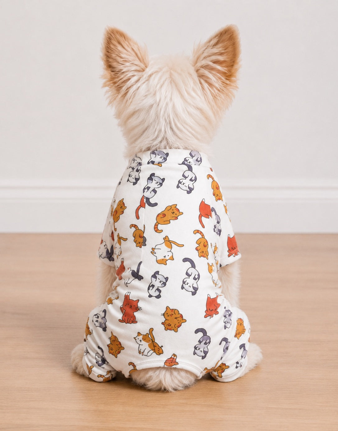 A small fluffy dog seen from behind, wearing a white Pajama romper covered in a colorful cartoon cat print, sitting on a light wood floor against a plain white wall.