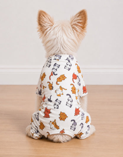 A small fluffy dog seen from behind, wearing a white Pajama romper covered in a colorful cartoon cat print, sitting on a light wood floor against a plain white wall.