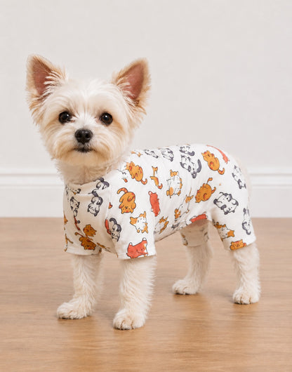 A small white dog standing on a wooden floor against a plain light wall, wearing a fitted short-sleeve Pagama Onesie with an allover cartoon animal print in orange, black, and gray on a white base.