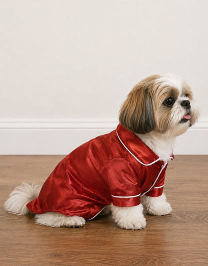 Side view of a small fluffy dog wearing shiny red Pajama Shirt with white trim, sitting on a wooden floor against a plain light wall.