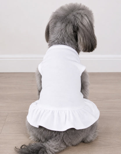 A gray dog seen from behind wearing a white t-shirt dress with a ruffled hem, sitting on a light wood floor against a white wall.