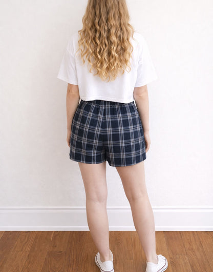Back view of woman wearing white tee with blue and white plaid Easy Elasticized Waist Shorts and white shoes with wooden floor and plain background.
