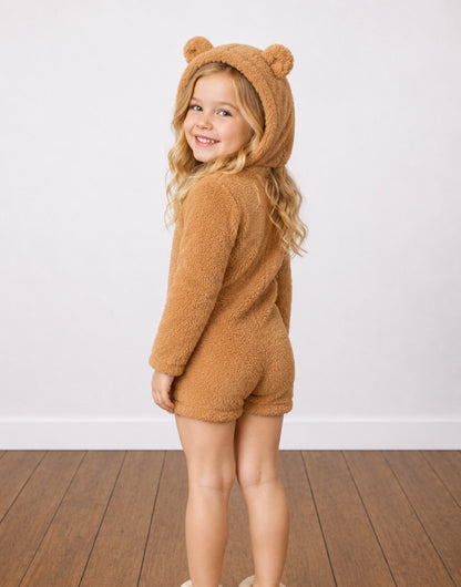 Back view of a young girl with blonde hair wearing a brown teddy fleece Hooded Romper with bear ears, standing on a wooden floor against a white wall.