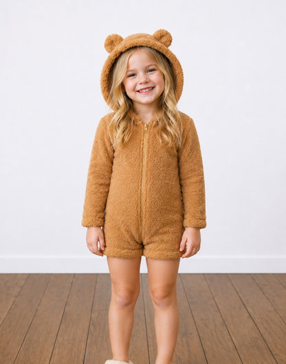 A young girl smiling while wearing a brown teddy fleece bear-themed Hooded Romper with ear details, standing on a wooden floor against a white wall.