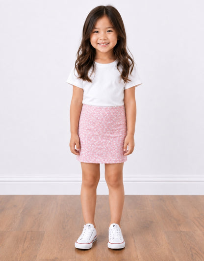 A young girl with long brown hair stands smiling, wearing a white short-sleeve t-shirt, a pink patterned Bodycon Mini Skirt, and white sneakers, set against a plain white wall and light wood floor.