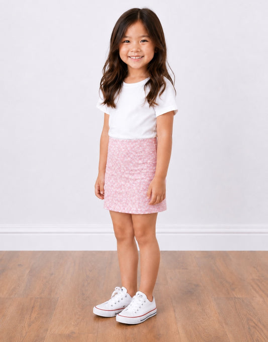Side view of a young girl with long brown hair stands smiling, wearing a white short-sleeve t-shirt, a pink patterned Bodycon Mini Skirt, and white sneakers, set against a plain white wall and light wood floor.