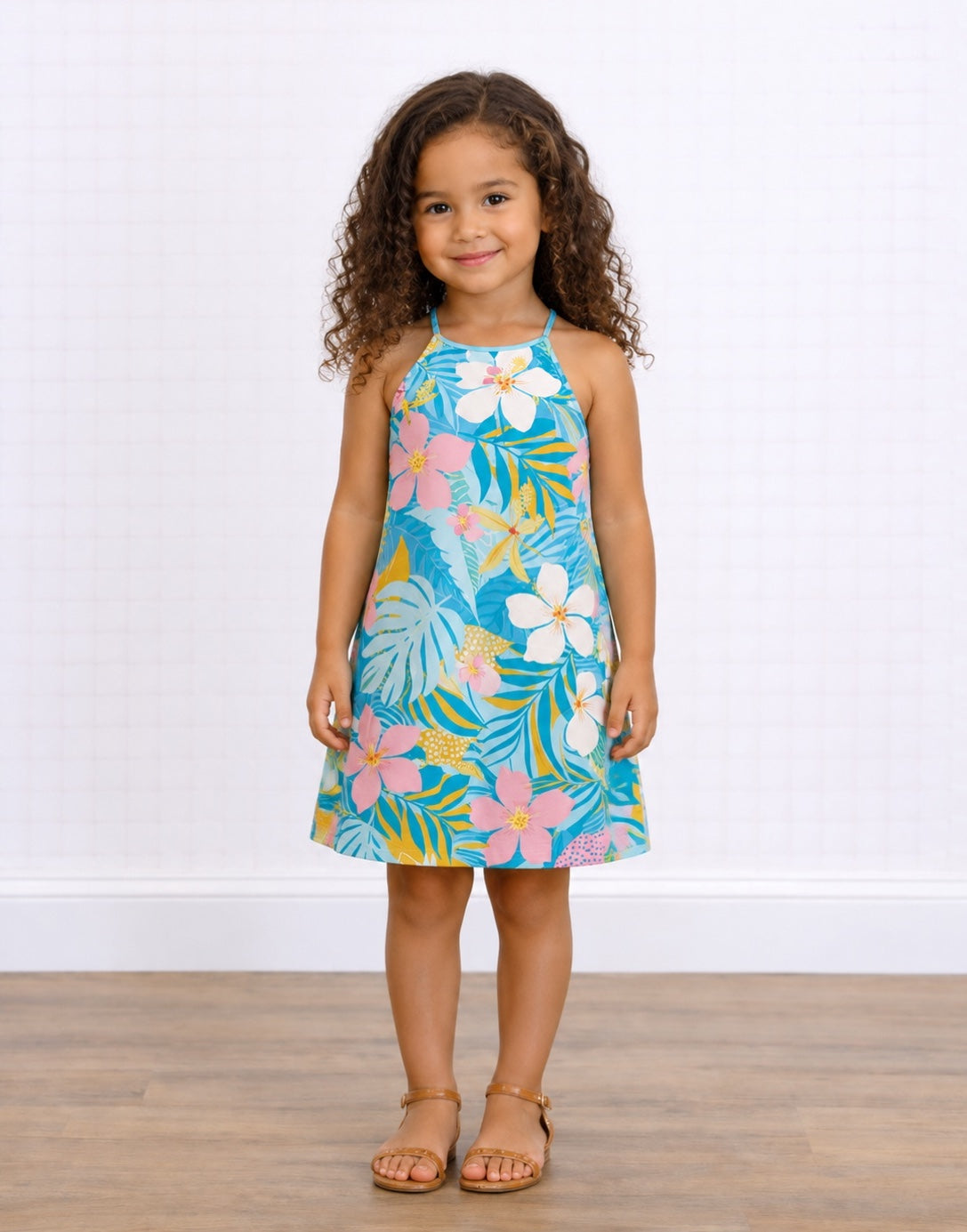 A young girl with curly brown hair stands wearing a Sleeveless Halter Cami Dress with a colorful tropical floral and leaf print on a turquoise background. She is wearing tan sandals and standing on a light wood floor against a white paneled wall.