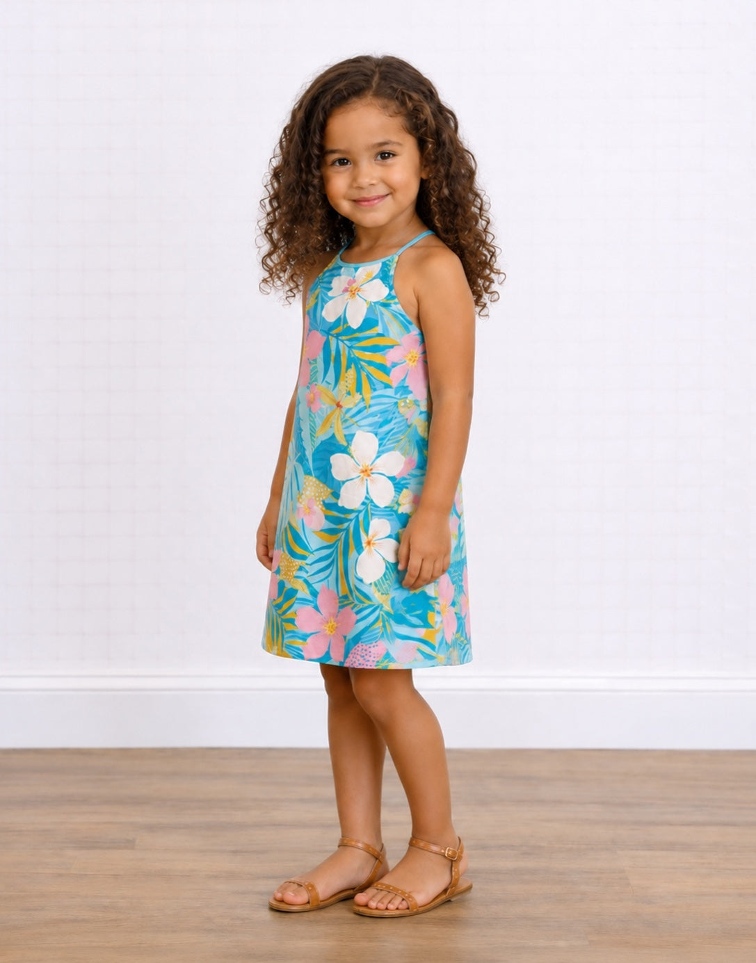 Side view of a young girl with curly brown hair stands wearing a Sleeveless Halter Cami Dress with a colorful tropical floral and leaf print on a turquoise background. She is wearing tan sandals and standing on a light wood floor against a white paneled wall.