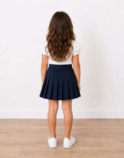 Back view of a young girl with long brown hair stands in a studio wearing a white short-sleeve puff-sleeve top and a navy blue High Waist Pleated Mini Skirt, paired with white sneakers, against a white wall and light wood floor.