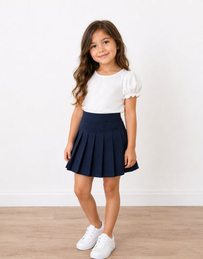 A young girl with long brown hair stands in a studio wearing a white short-sleeve puff-sleeve top and a navy blue High Waist Pleated Mini Skirt, paired with white sneakers, against a white wall and light wood floor.
