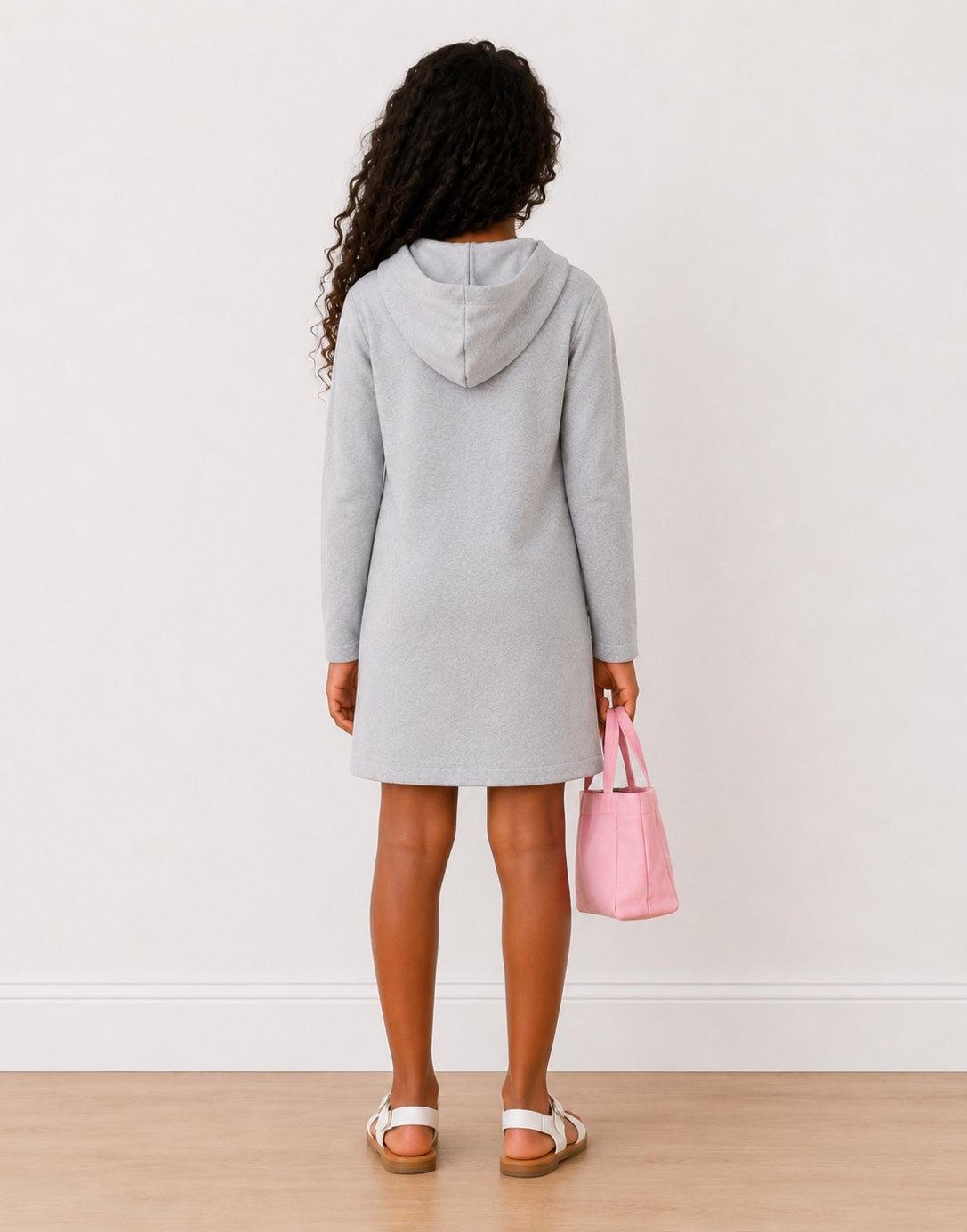 A girl shown from the back wearing a light gray Hooded Long Sleeve Sweatshirt dress, holding a pink handbag, standing on a light wood floor against a white wall.