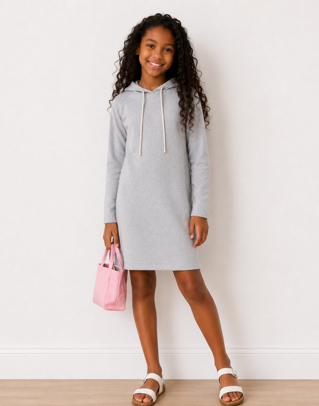 A girl wearing a light gray Hooded, Long Sleeve, Knit Sweatshirt Dress with drawstrings, holding a pink handbag, standing on a light wood floor against a white wall.