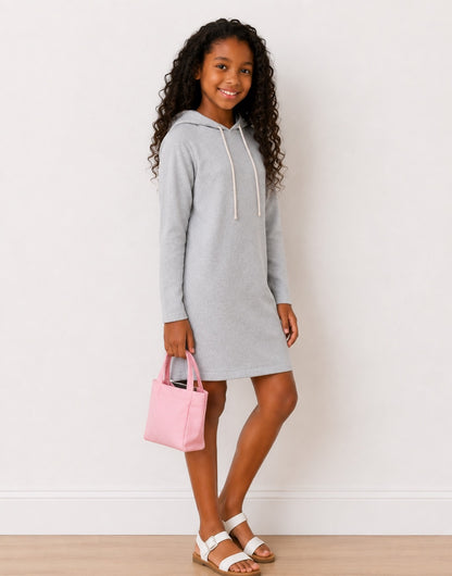 Side view of a girl wearing a light gray Hooded, Long Sleeve, Knit Sweatshirt Dress with drawstrings, holding a pink handbag, standing on a light wood floor against a white wall.