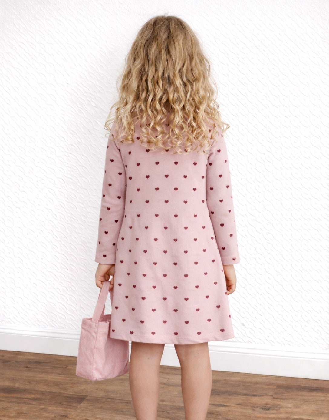 Back view of child wearing a pink dress with heart pattern holding a matching bag against a white wall.
