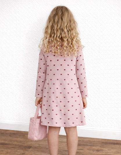 Back view of child wearing a pink dress with heart pattern holding a matching bag against a white wall.