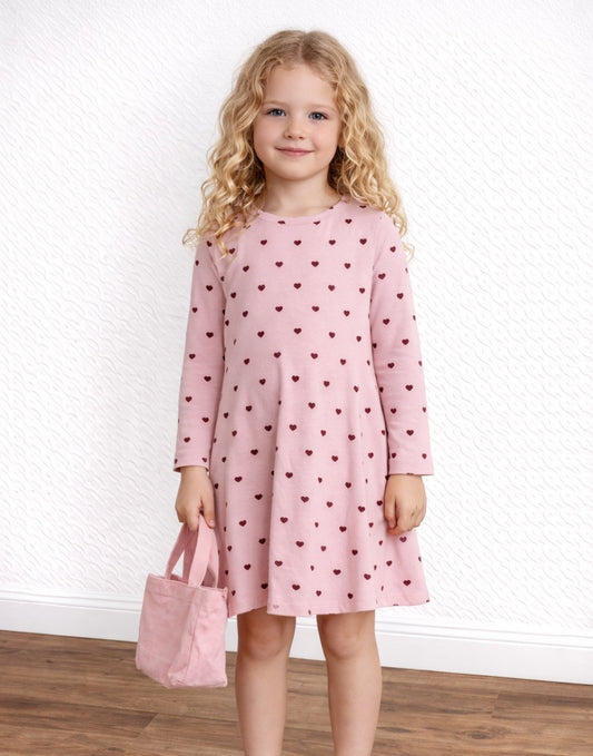 Child wearing a pink dress with heart pattern holding a matching bag against a white wall.