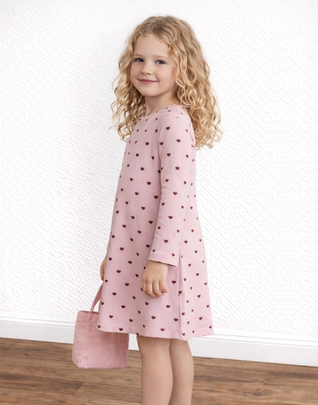 Side view of child wearing a pink dress with heart pattern holding a matching bag against a white wall.