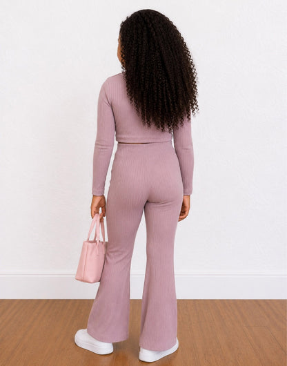 A young girl shown from behind wearing a matching dusty pink ribbed two-piece Loungewear Set consisting of a long-sleeve crop top and high-waisted flared pants, holding a small pink handbag on a wooden floor against a white wall.