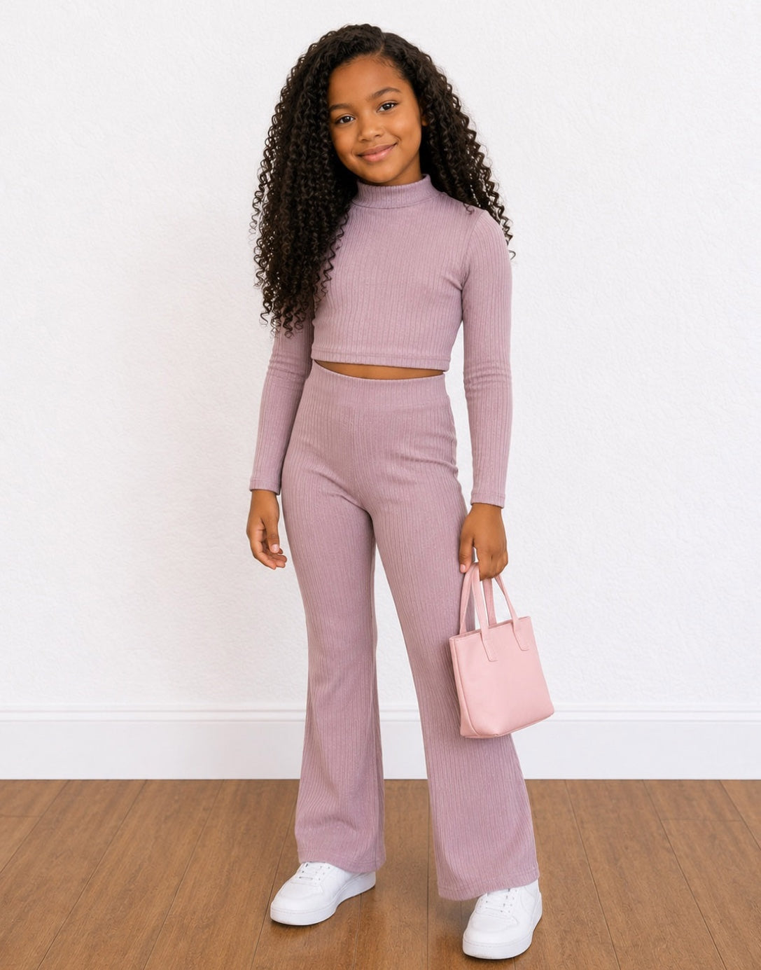 A young girl with long curly black hair wearing a matching ribbed two-piece Loungewear Set in dusty mauve, consisting of a long-sleeve crop top and high-waisted wide-leg pants. She is holding a small light pink tote bag and wearing white sneakers, set against a white wall and wood floor.