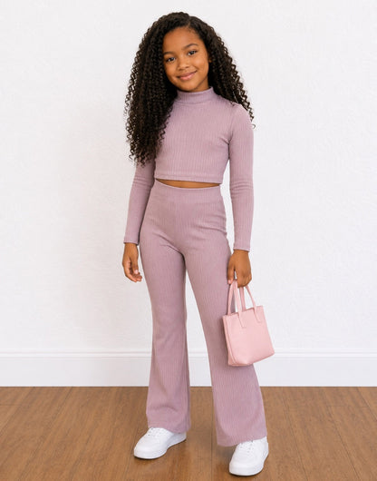 A young girl with long curly black hair wearing a matching ribbed two-piece Loungewear Set in dusty mauve, consisting of a long-sleeve crop top and high-waisted wide-leg pants. She is holding a small light pink tote bag and wearing white sneakers, set against a white wall and wood floor.