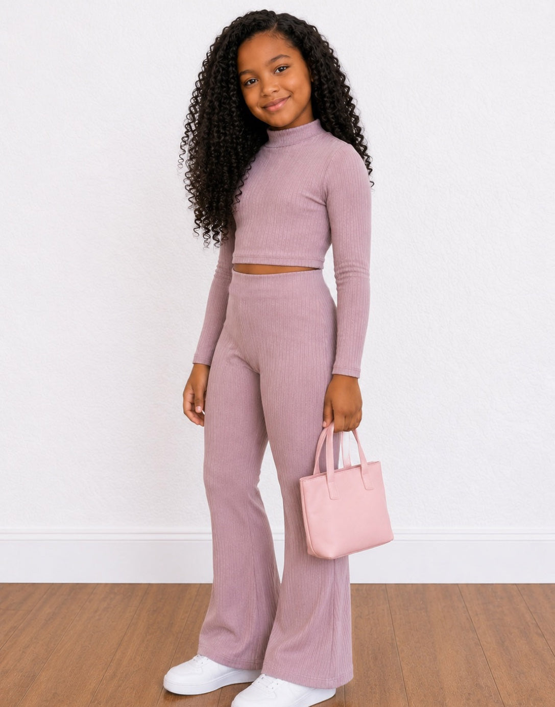 Side view of a young girl with long curly black hair wearing a matching ribbed two-piece Loungewear Set in dusty mauve, consisting of a long-sleeve crop top and high-waisted wide-leg pants. She is holding a small light pink tote bag and wearing white sneakers, set against a white wall and wood floor.