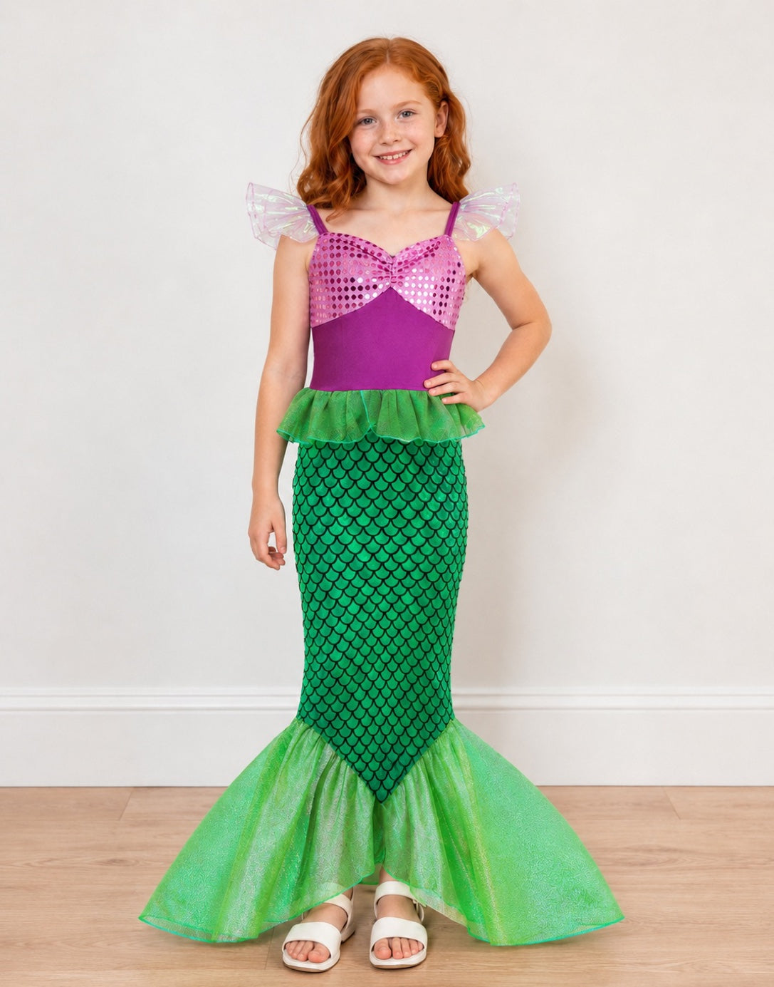 A young girl with red hair wearing a Mermaid Costume, featuring a purple sequined top with pink ruffled shoulder details, a green ruffle waist, and a green scale-patterned mermaid tail flared out, standing against a white wall and light wood floor.