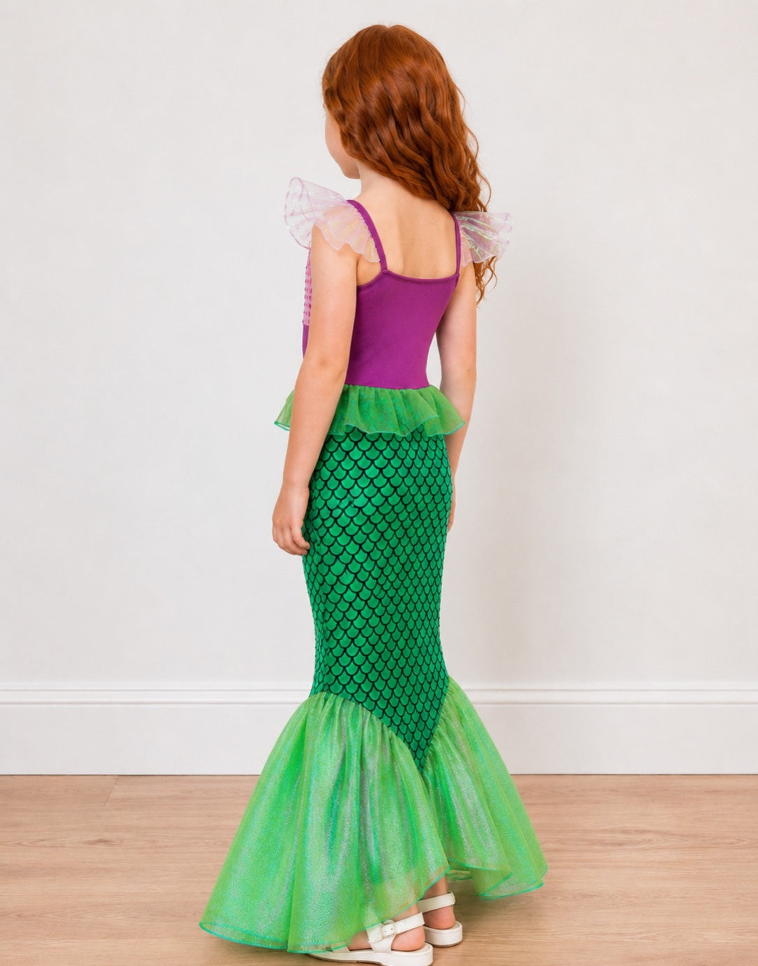 A young girl shown from the side wearing a mermaid costume with a purple top, green ruffled trim, and a green scale-patterned mermaid tail on a light wood floor against a white wall.