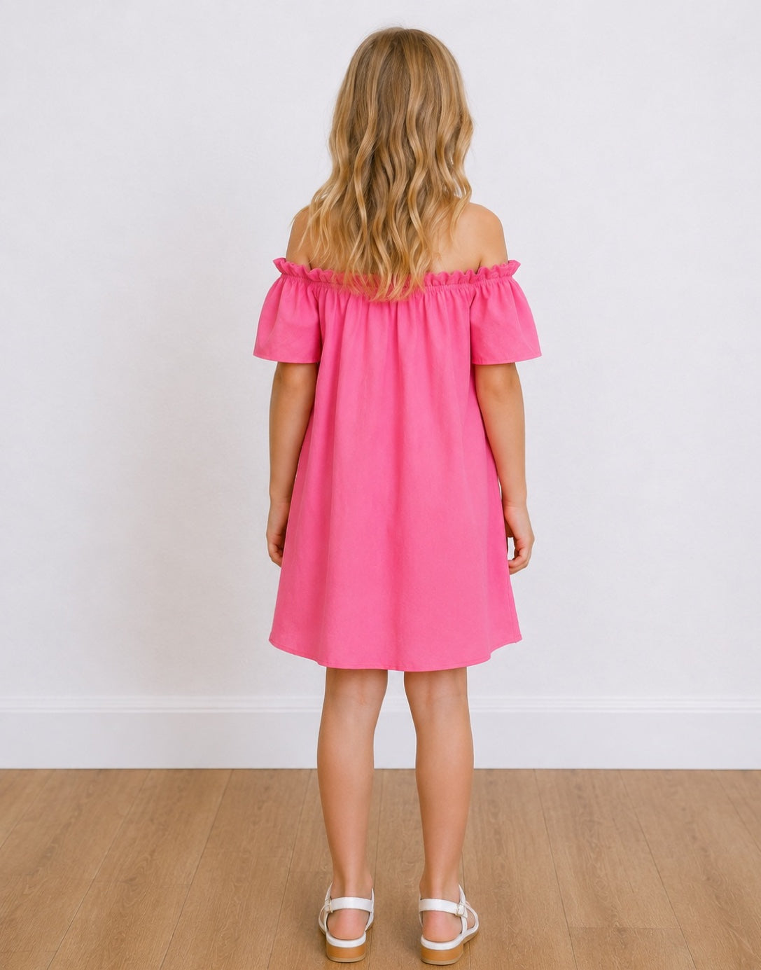 Back view of a young girl with blonde hair stands smiling, wearing a bright pink Off Shoulder Short-Sleeve Dress and white sandals, set against a white wall and light wood floor.
